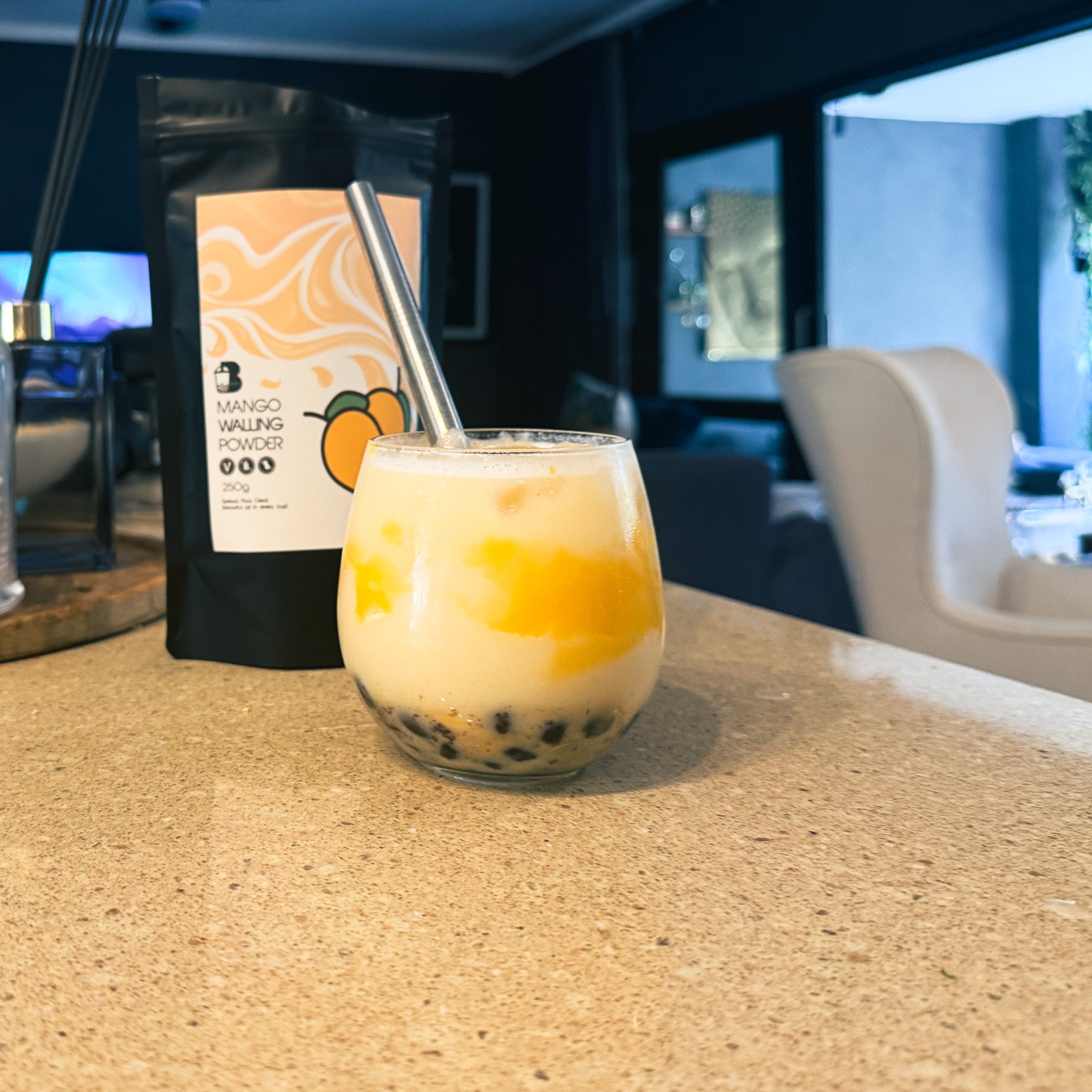 Glass of a layered drink with a straw on a countertop, with a package of mango powder in the background.